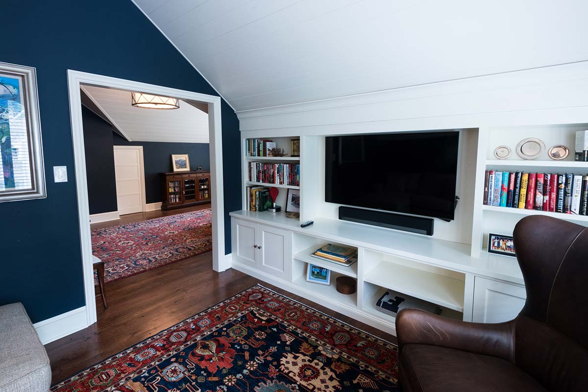 Cozy loft room with a dark blue vaulted ceiling, white built-in shelves, a large TV, and a leather chair on a vibrant rug.