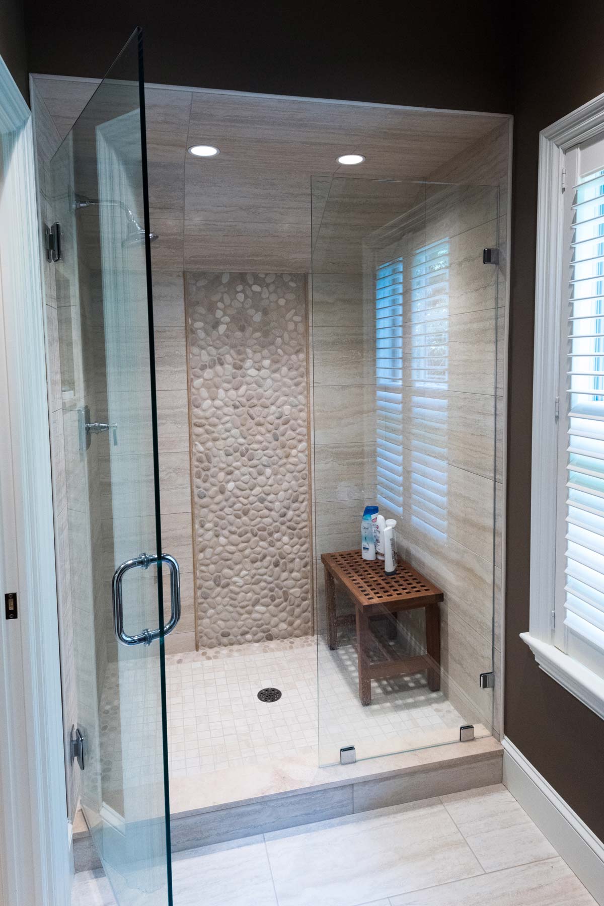 Luxurious bathroom with a glass-enclosed shower, pebble wall, and wooden bench, featuring natural light from large windows.