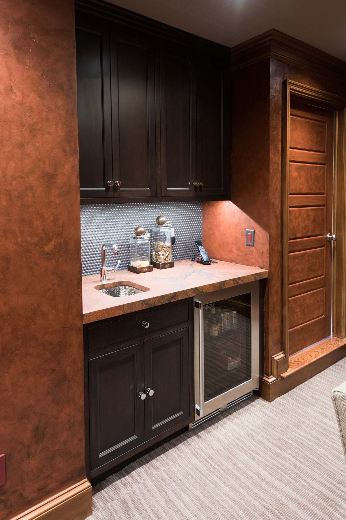 Stylish wet bar with dark cabinets, a textured backsplash, and a built-in wine cooler, set against a warm terracotta wall.