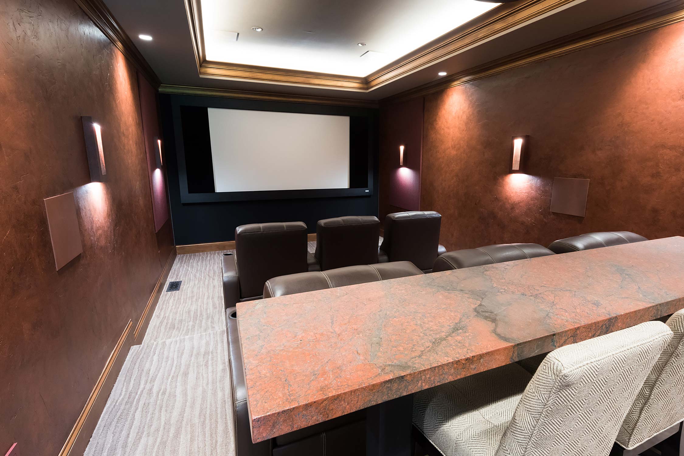 Luxurious home theater with leather seating, large screen, and stone bar counter, illuminated by wall sconces and recessed lighting.