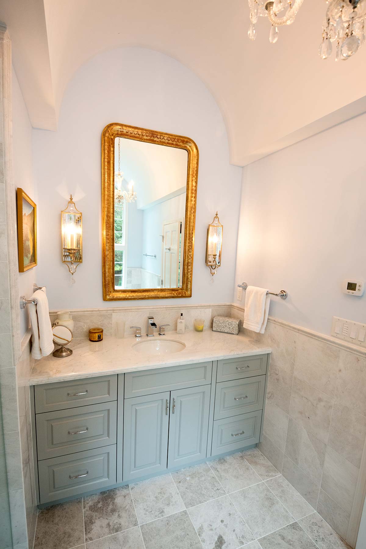 Elegant bathroom with arched ceiling, crystal chandelier, and large gold mirror. Features marble vanity, gray cabinets, tiled floors, and ornate wall sconces.