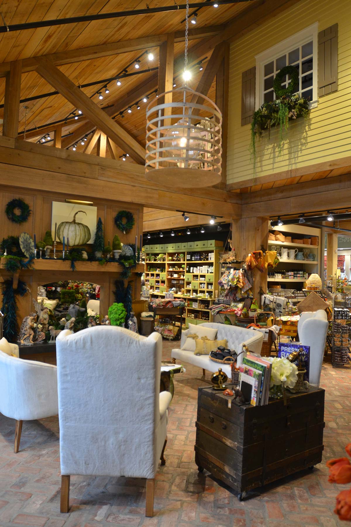 Cozy store interior with brick floor, white armchairs, wooden beams, and shelves of decor items. A chandelier hangs from the ceiling, and wreaths adorn the walls.