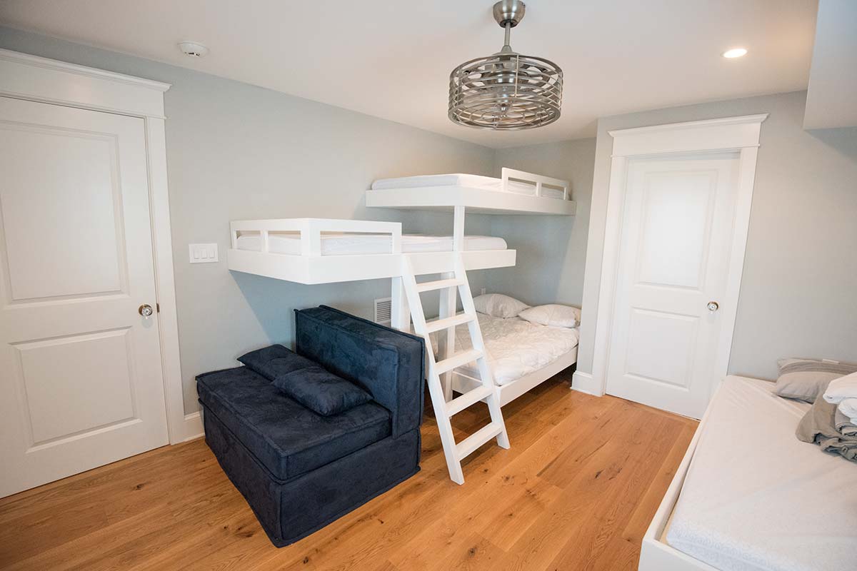 A small bedroom with white bunk beds, a blue sofa, a single bed, wooden floors, and a ceiling fan.