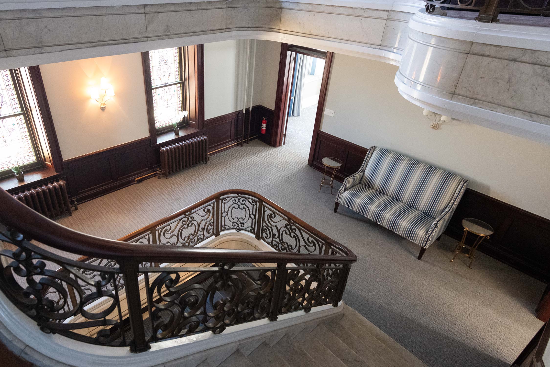 A view from above of a marble staircase with ornate black railings, leading to a landing with a striped blue sofa, small tables, and stained glass windows.