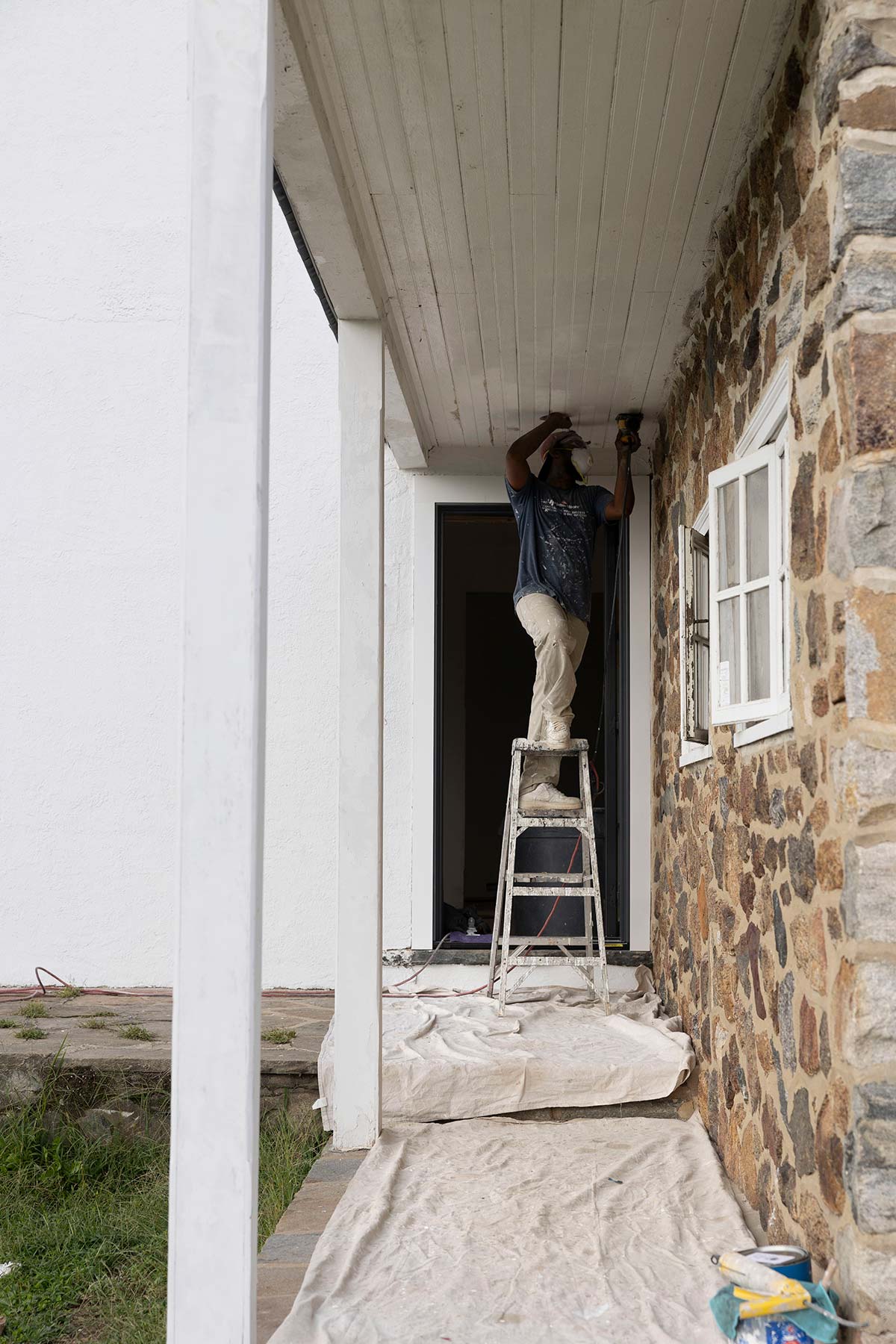Worker on a ladder painting the ceiling of a porch with stone walls, white columns, and a window, with drop cloths and paint cans below.