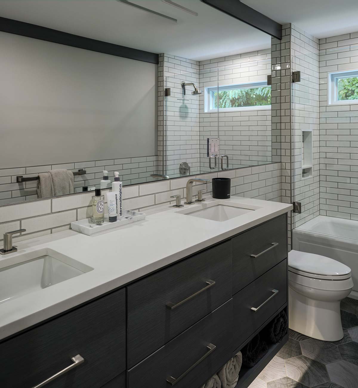 Modern bathroom with dark gray vanity, double sinks, and white subway tile walls. A large mirror, glass shower, and white tub are visible, with a hexagonal tile floor.