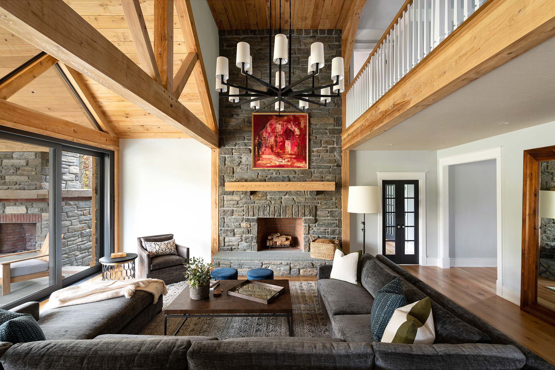 Spacious living room with stone fireplace, wooden beams, large chandelier, gray sectional sofa, and a loft balcony with hardwood floors.