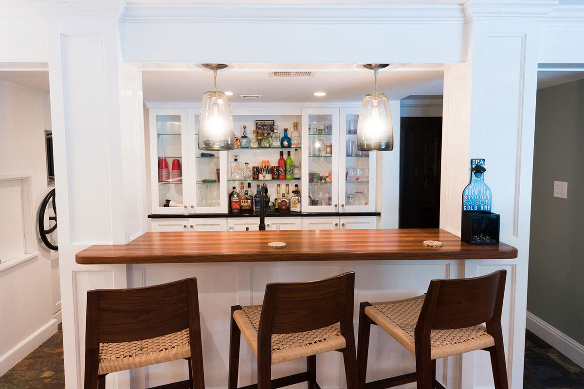 Chic bar area with a wooden counter, modern stools, and glass shelves stocked with bottles. Features warm lighting and a cozy, stylish ambiance.