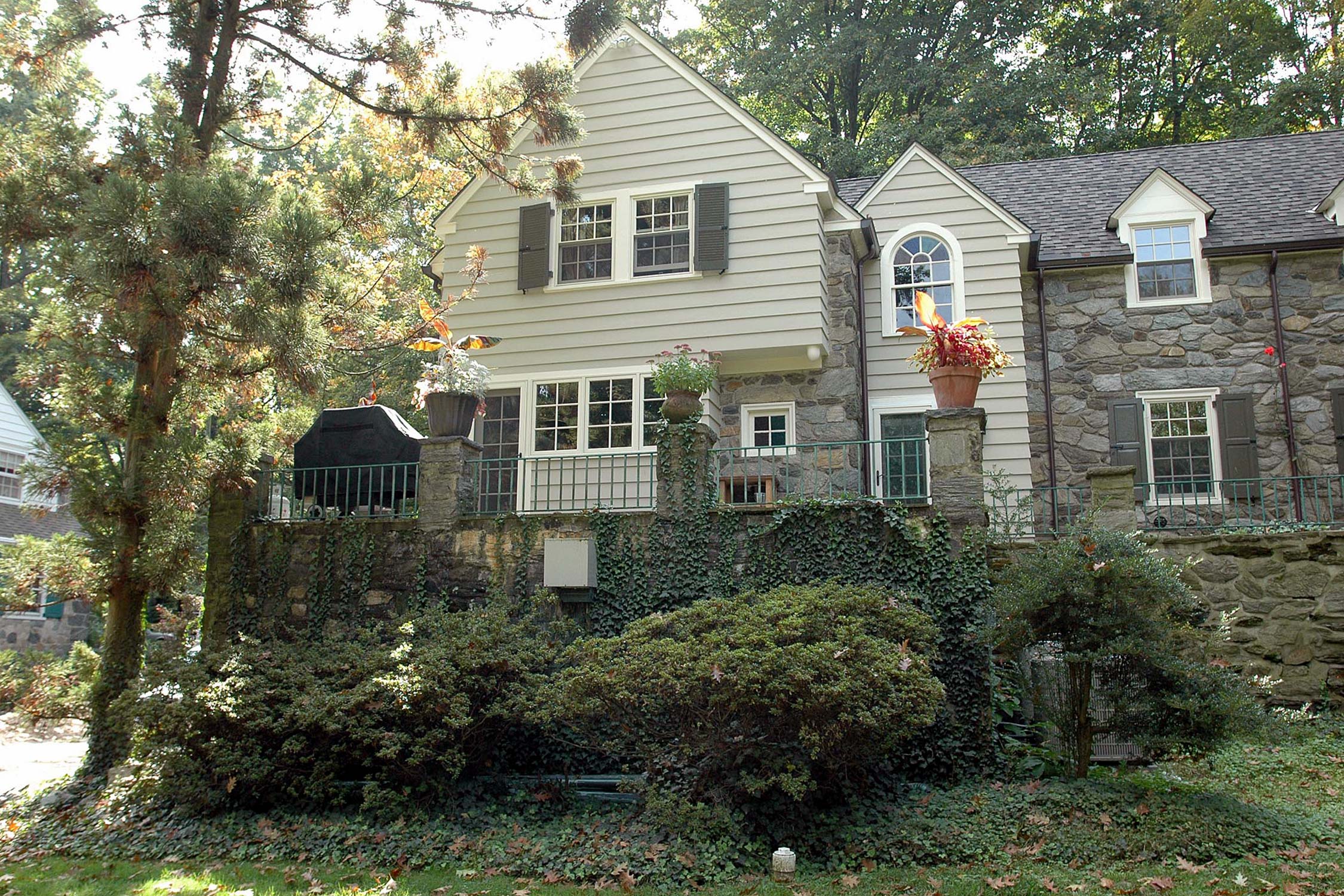 Charming two-story home with stone and siding exterior, surrounded by lush greenery and vibrant fall foliage. Features include a balcony and potted flowers.