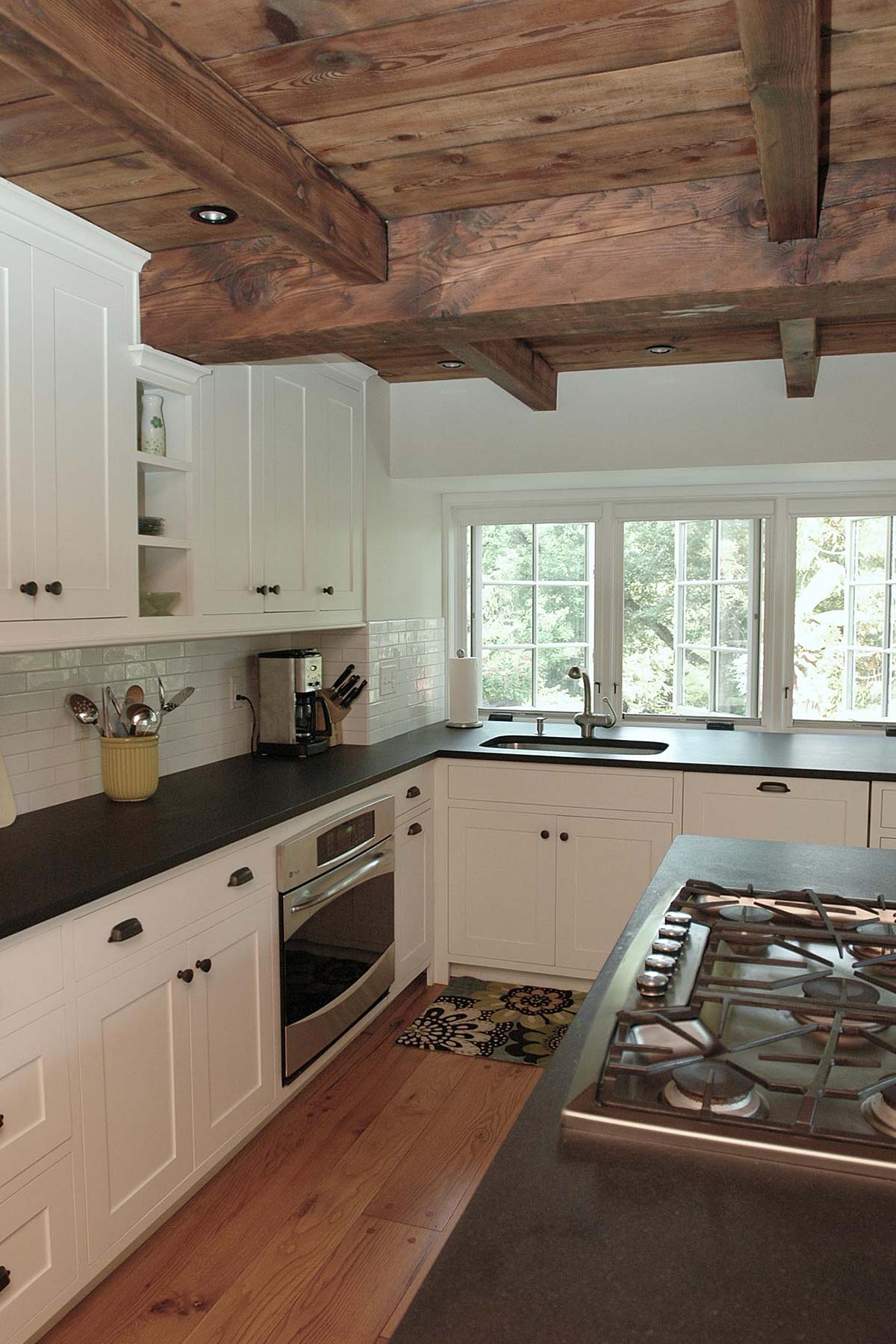 Modern kitchen with wooden beams, white cabinets, black countertops, and large windows. Features a gas stove, oven, and cozy hardwood floors.