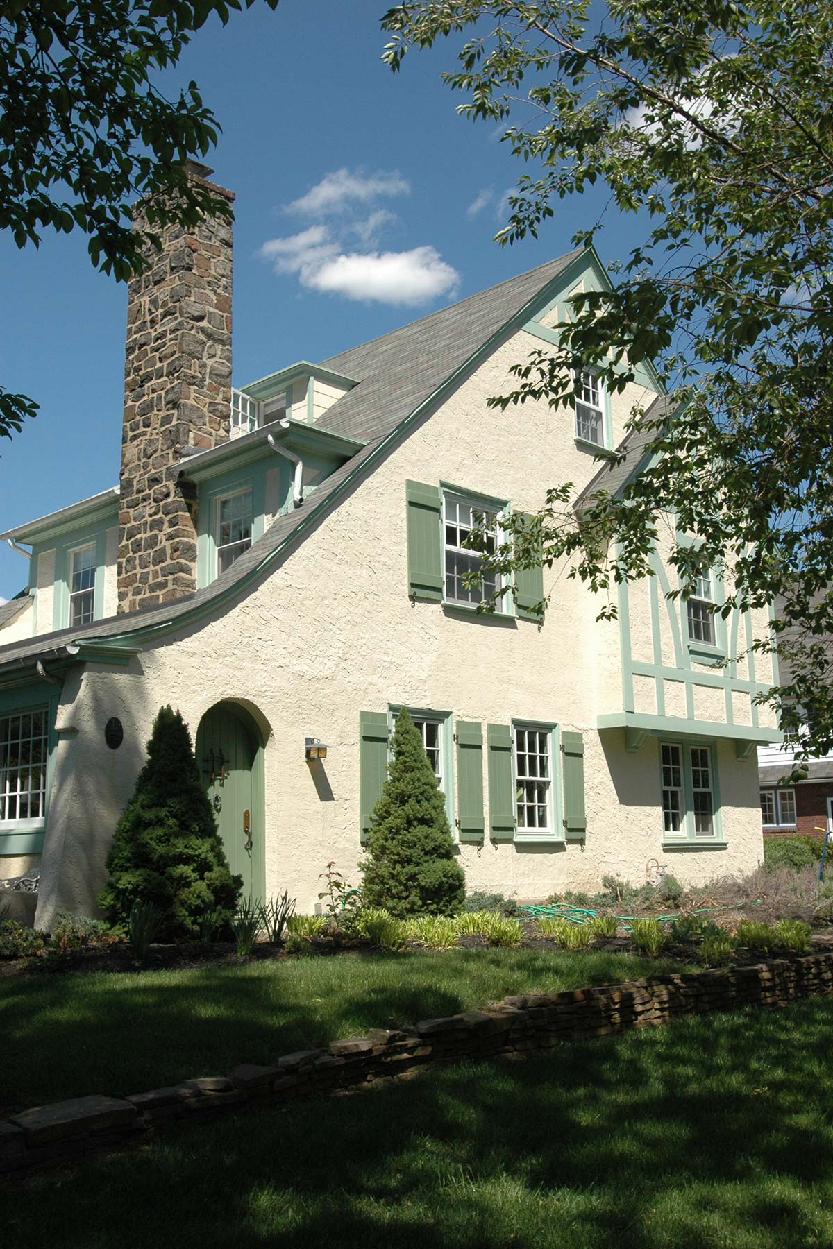 Quaint home with stucco exterior, green shutters, and stone chimney. Surrounded by lush greenery and a stone wall under a bright blue sky.