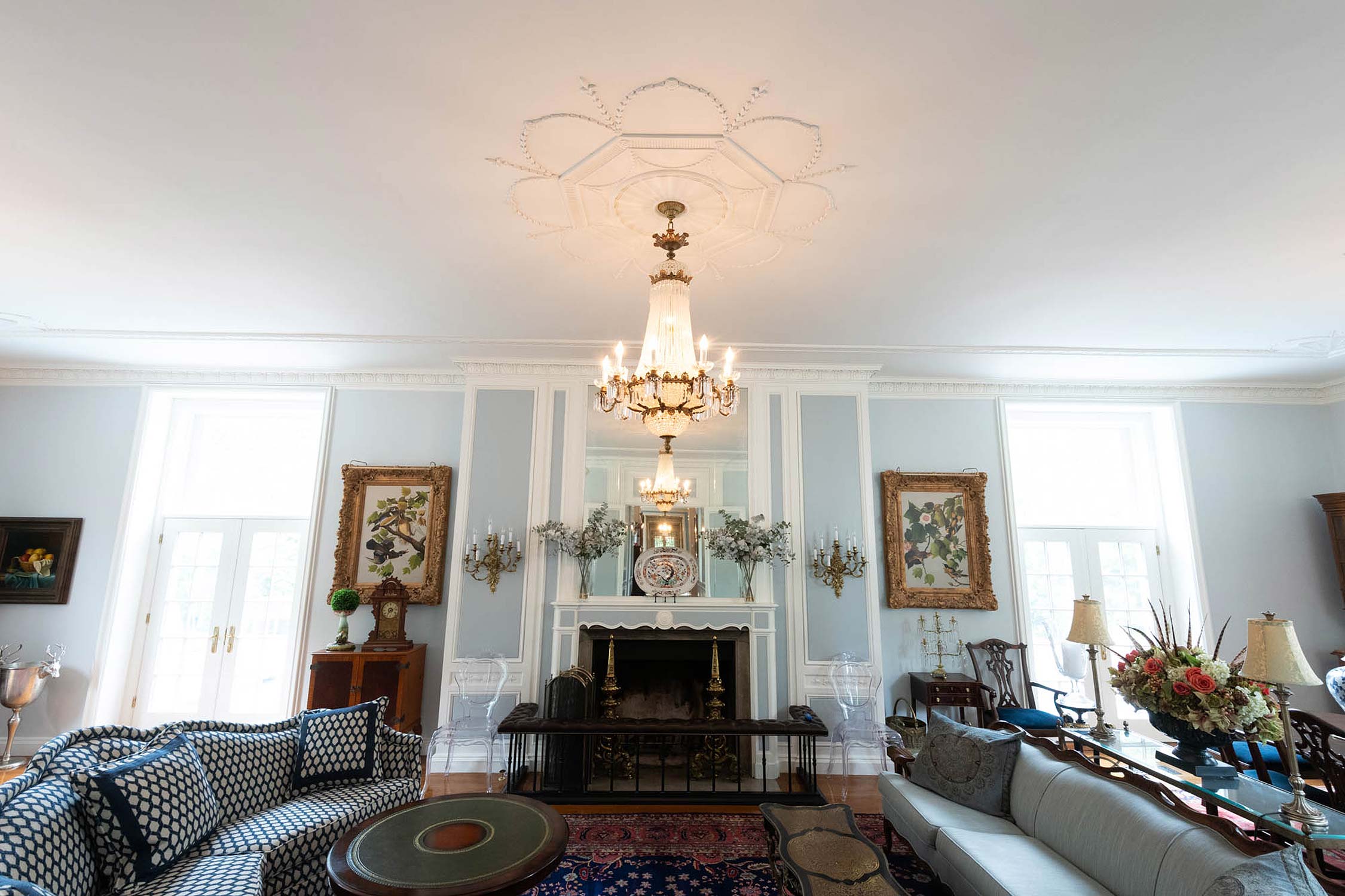 Elegant living room with ornate chandelier, fireplace, and classic decor. Features sofas, artwork, and natural light from large windows.