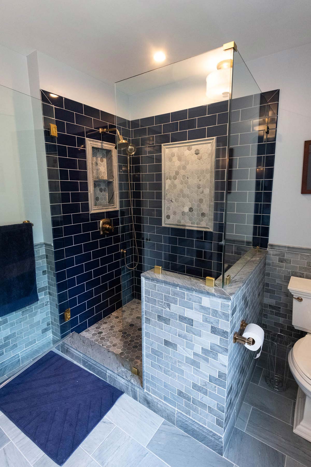 Modern bathroom with glass shower featuring blue subway tiles, hexagonal mosaic, and brass fixtures. Includes toilet and towel.