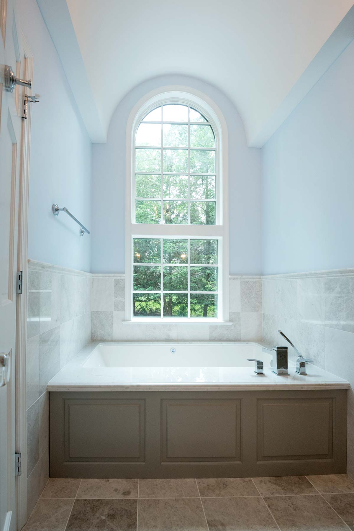 Serene bathtub under arched window with garden view. Features marble surround, gray paneling, tiled floors, chrome fixtures, and natural light.