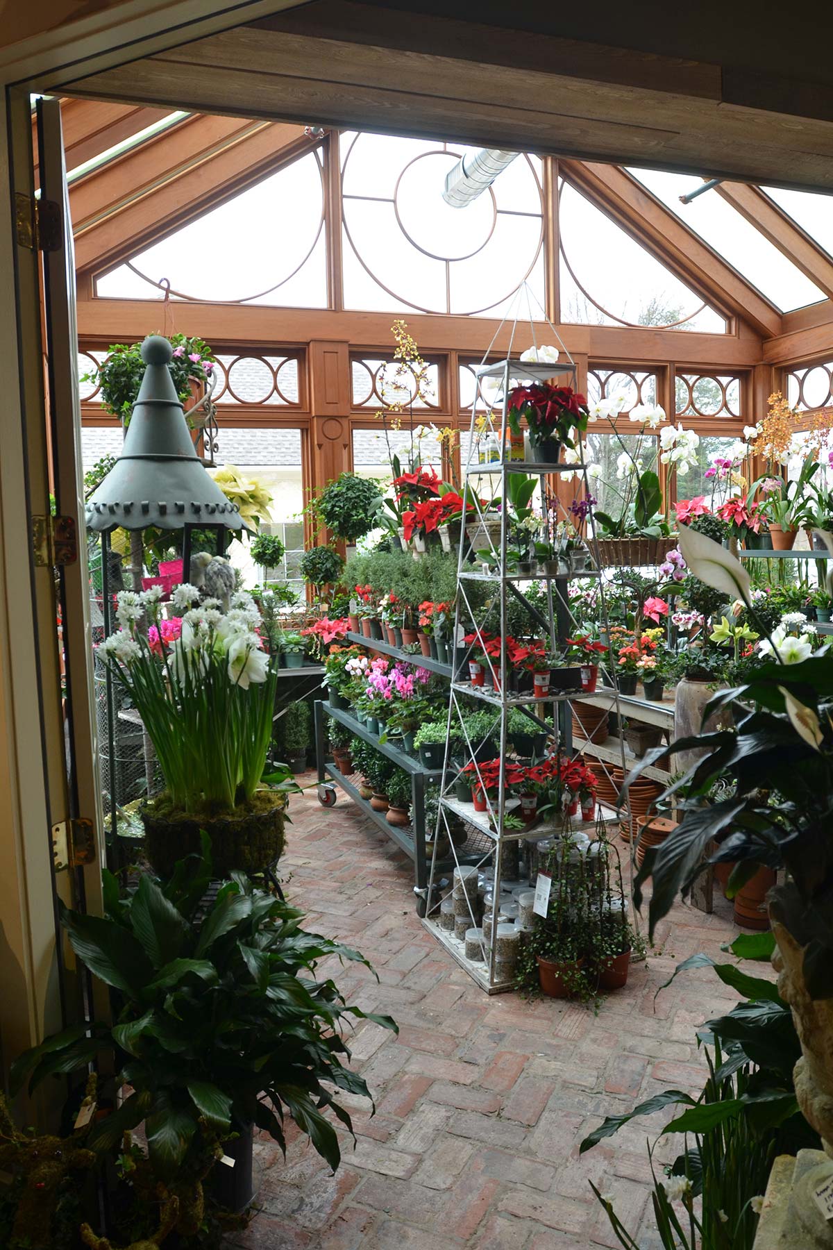 Greenhouse with brick floor, filled with colorful flowers on shelves, in pots, and hanging baskets. Wooden frame and glass walls let in natural light.