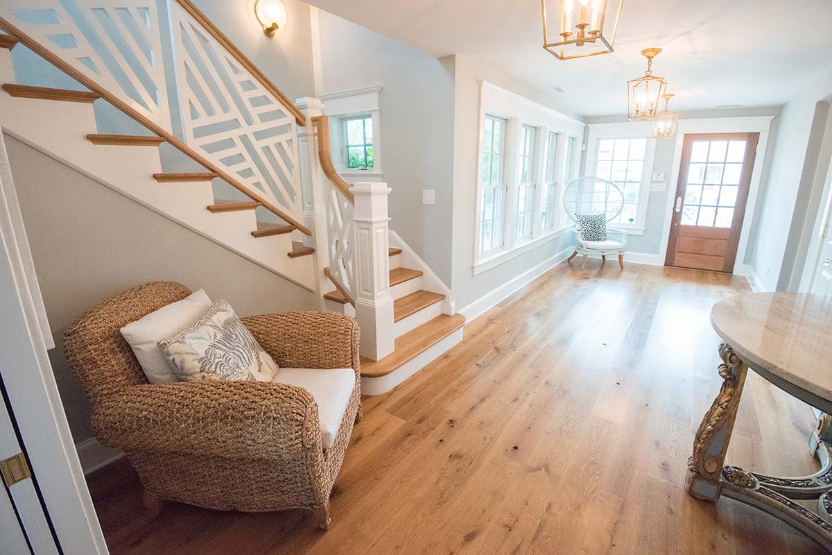A bright entryway with a wooden staircase, wicker chair, marble console table, lantern lights, and large windows.