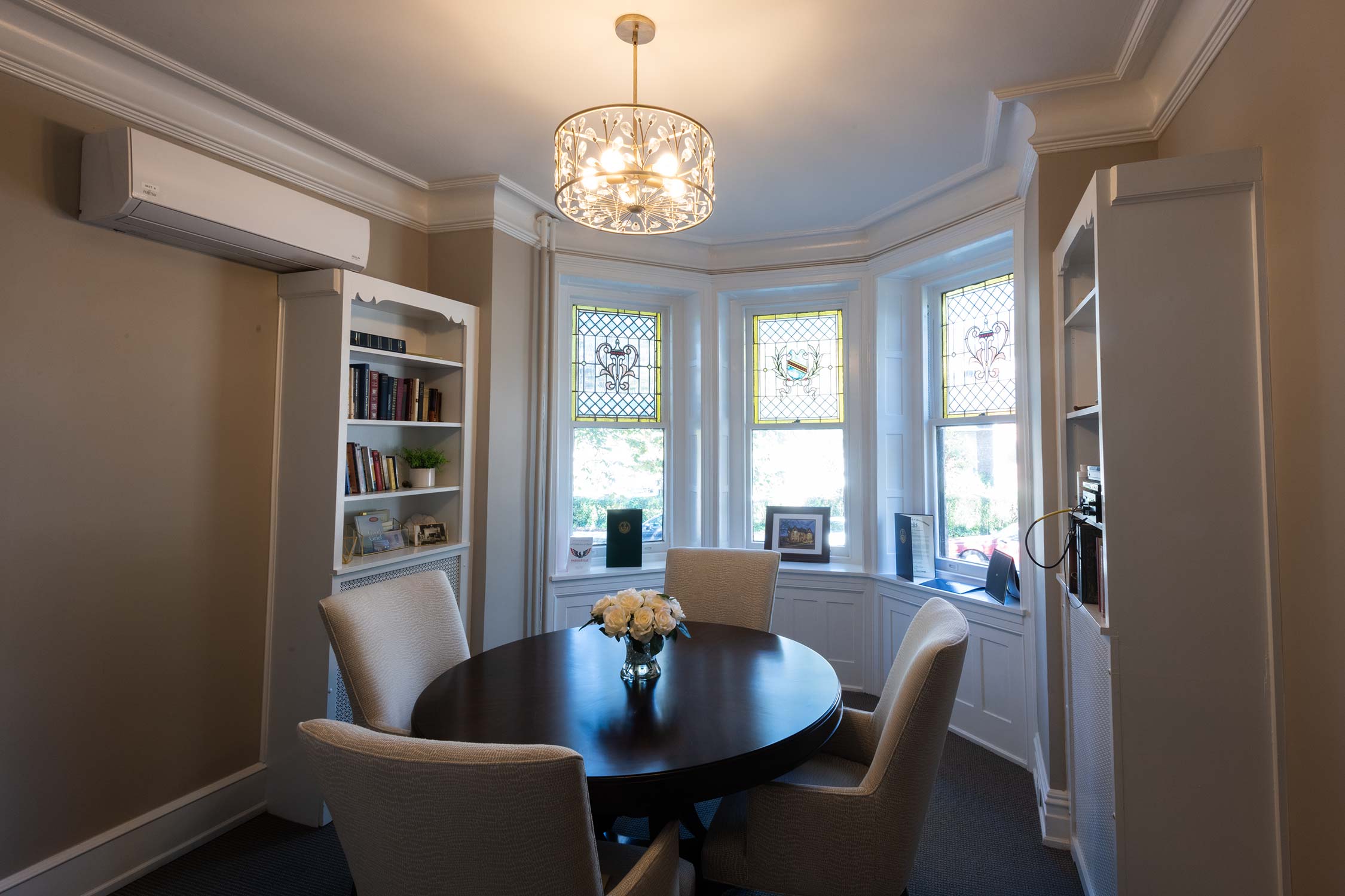 A cozy room with a round table, beige chairs, and a vase of white roses. Stained glass windows, bookshelves, and a chandelier add elegance, with a laptop on a shelf.