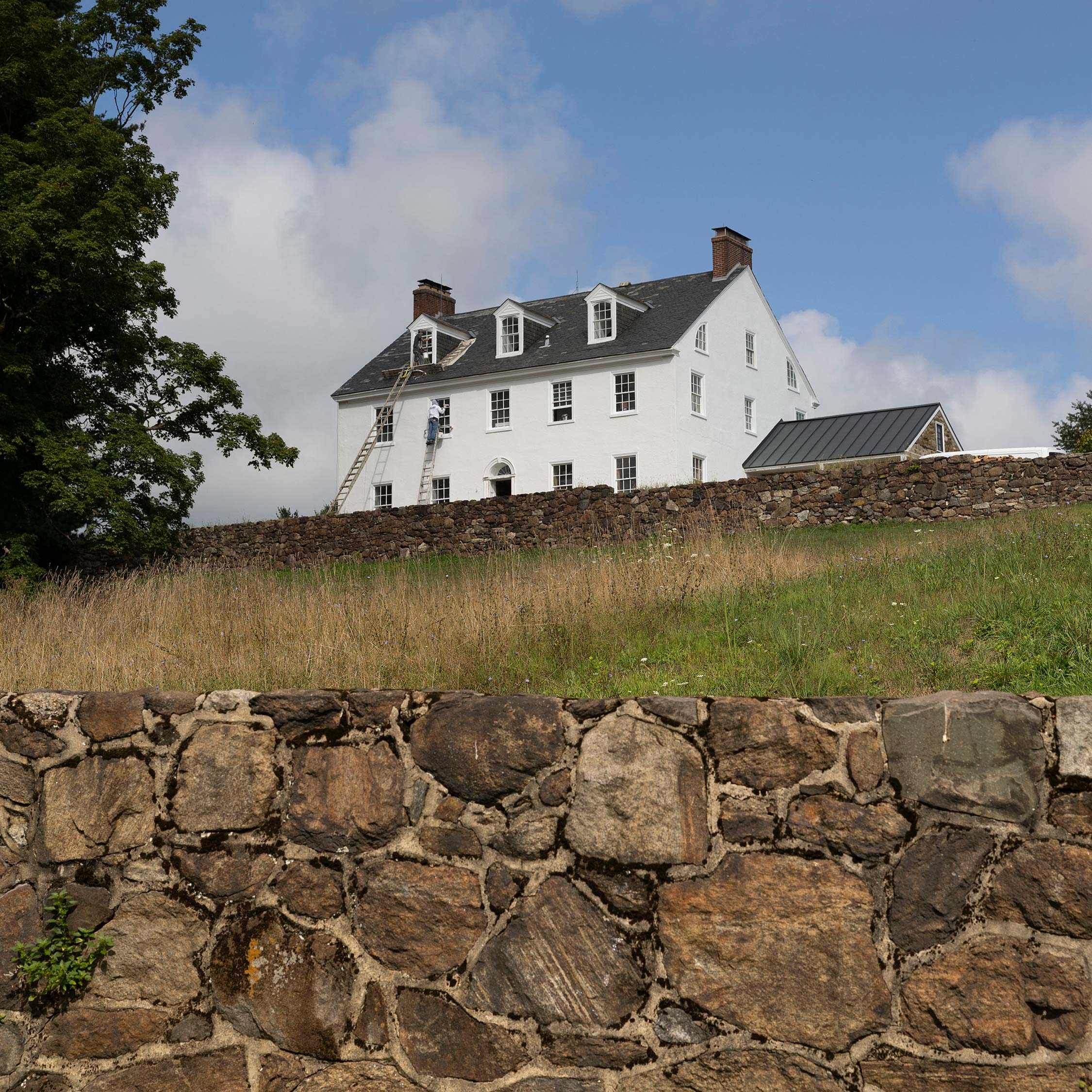 Worker on ladder painting a white historic home with dormer windows and stone wall, surrounded by grass and trees under a partly cloudy sky.
