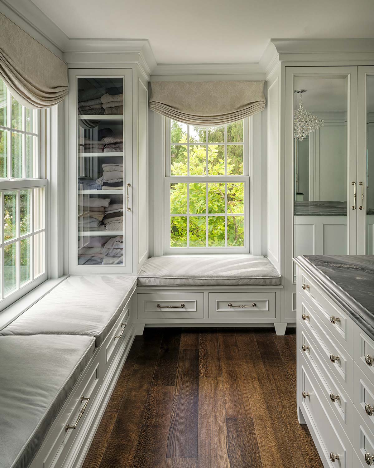 Bright dressing room with white cabinets, cushioned window seat, large windows with greenery view, and a marble-top island.