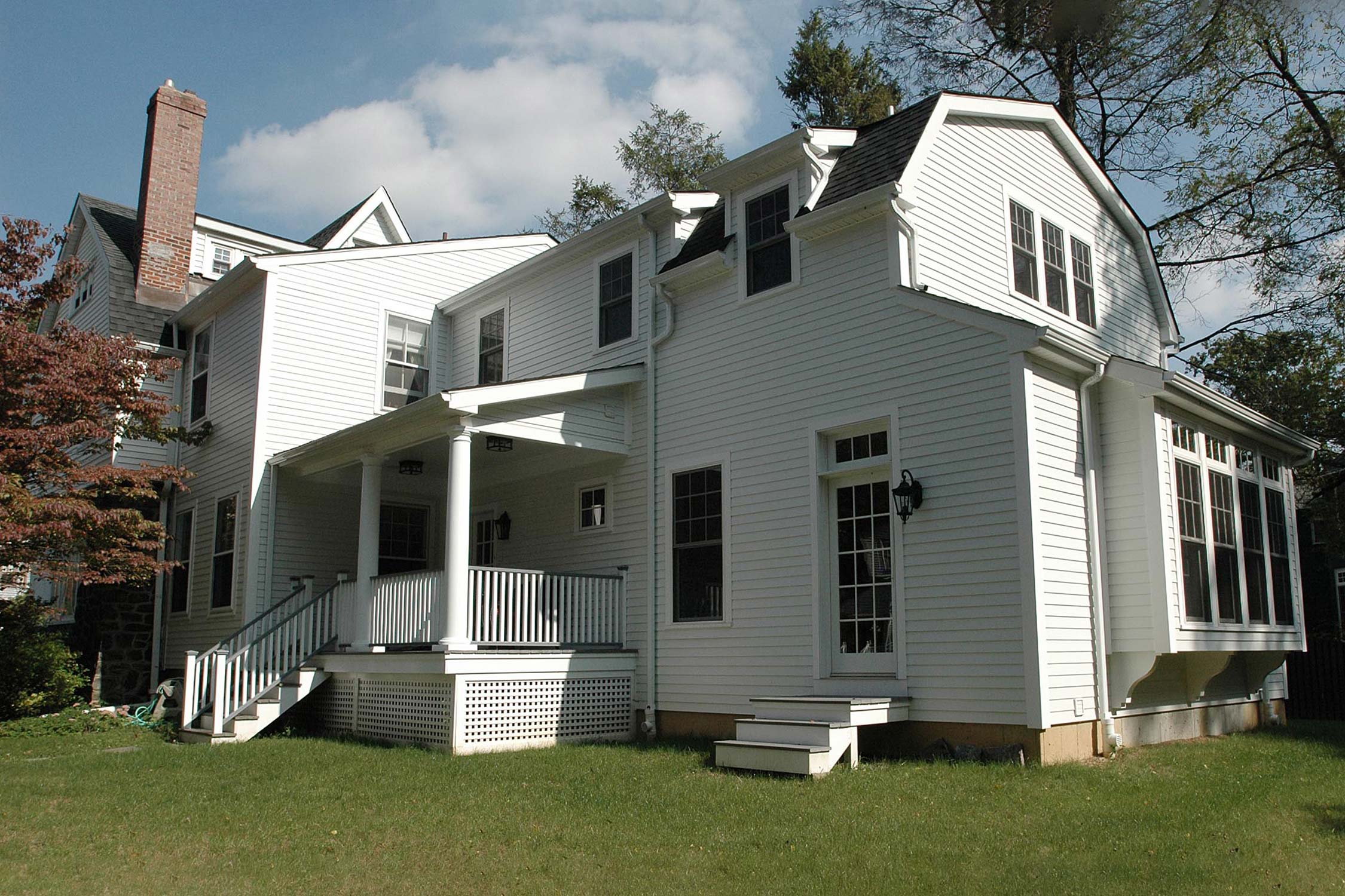 Classic white house with porch, brick chimney, and large windows, surrounded by green lawn and trees under a partly cloudy sky.