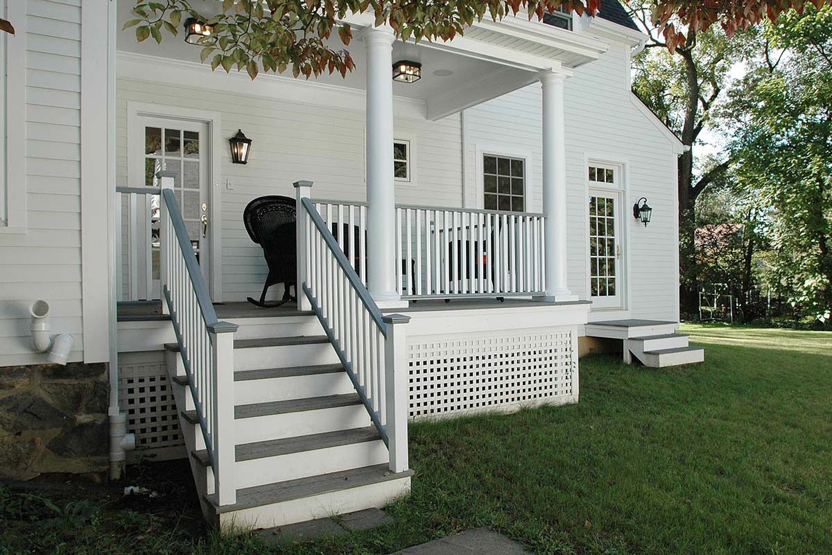 Charming white porch with columns, stairs, rocking chair, and lanterns on a classic house, surrounded by green lawn and trees.