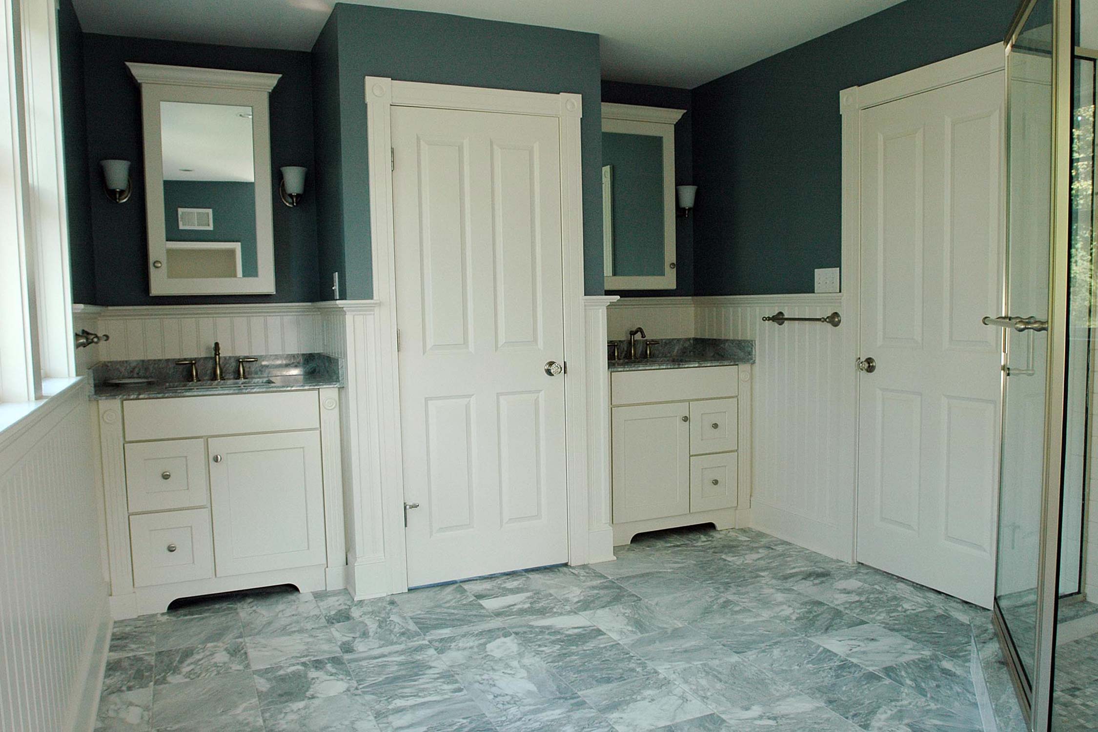 Elegant bathroom with dual vanities, marble flooring, teal walls, and glass shower. Features mirrors and wainscoting.