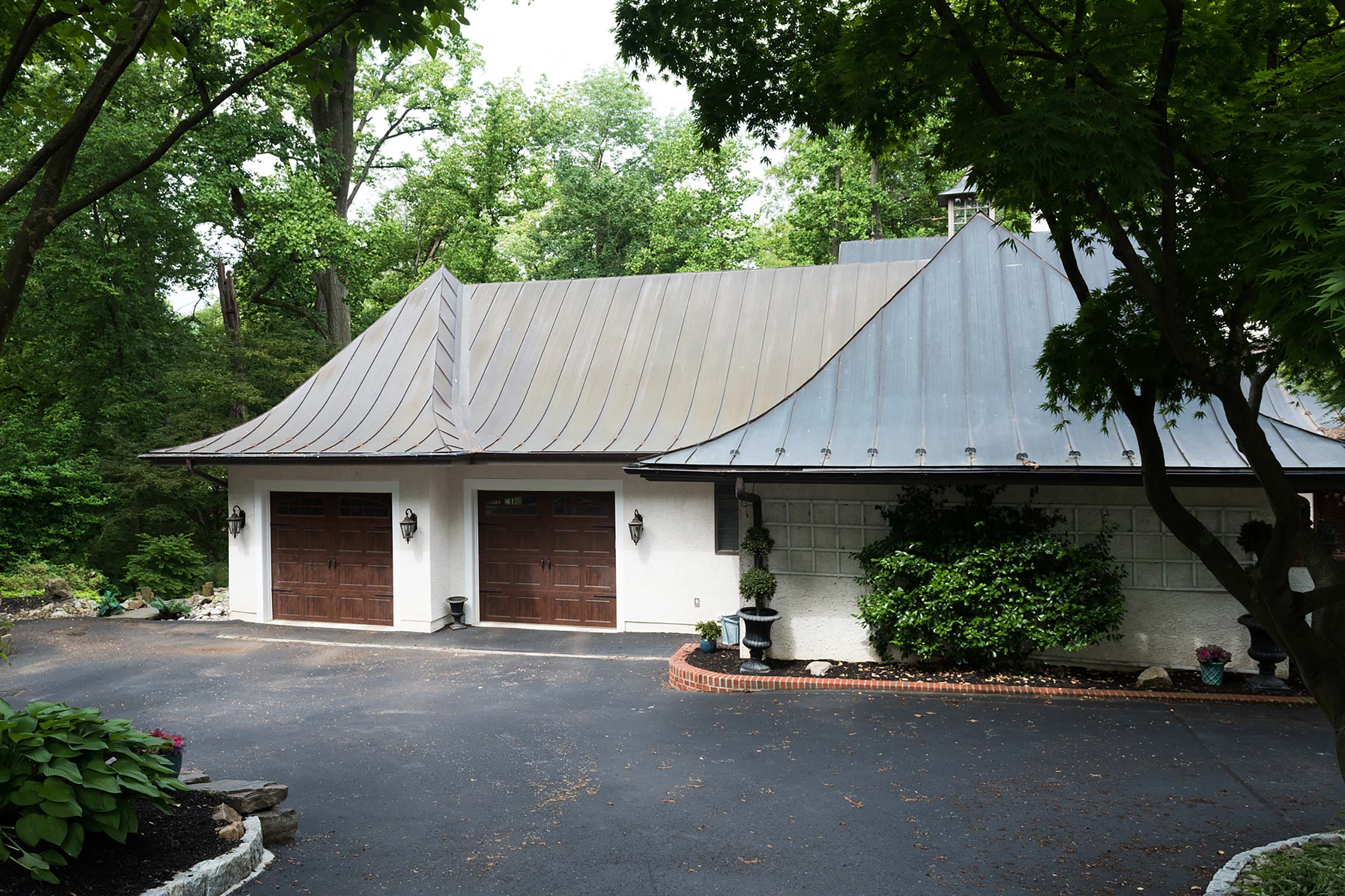 Modern garage with a sleek metal roof, double wooden doors, and landscaped surroundings, nestled among lush green trees.