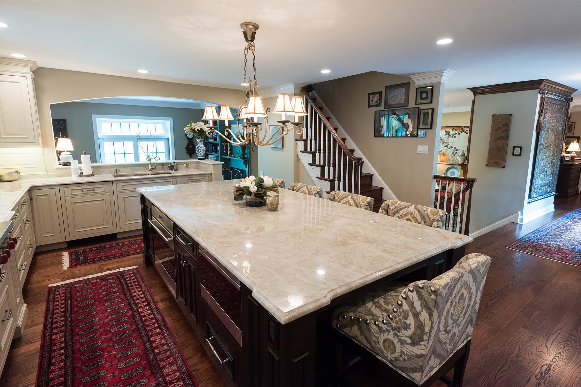 Spacious kitchen with a large marble island, chandelier, and upholstered chairs. Features a staircase, rugs, and natural light.
