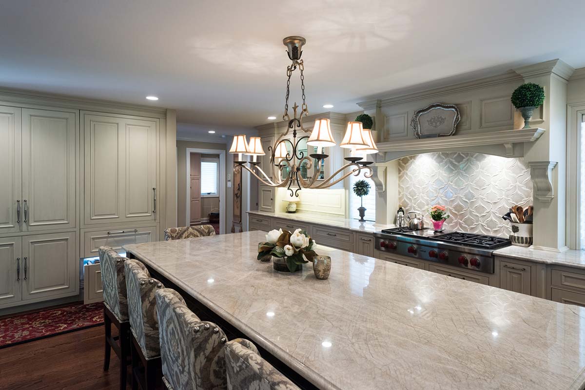 Elegant kitchen with a large marble island, chandelier, and upholstered chairs. Features a gas stove and decorative backsplash.