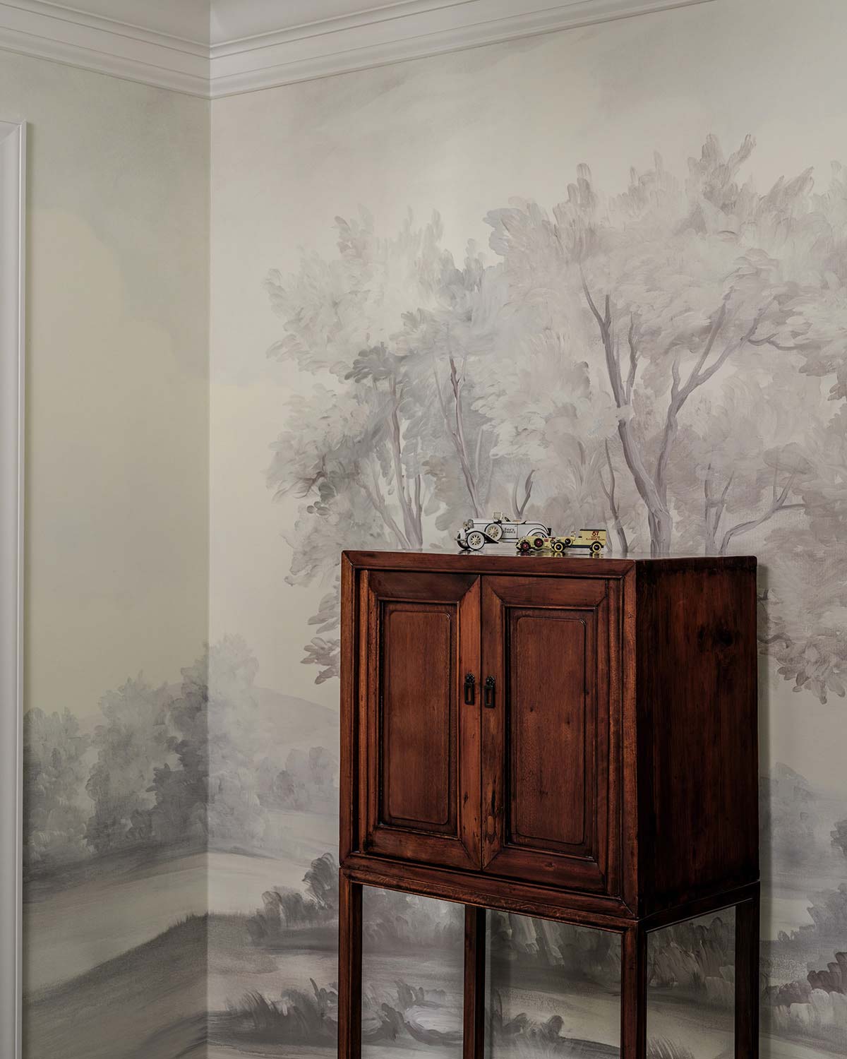 Cozy room with a dark wooden cabinet, toy cars on top, and walls featuring a subtle painted forest landscape mural.
