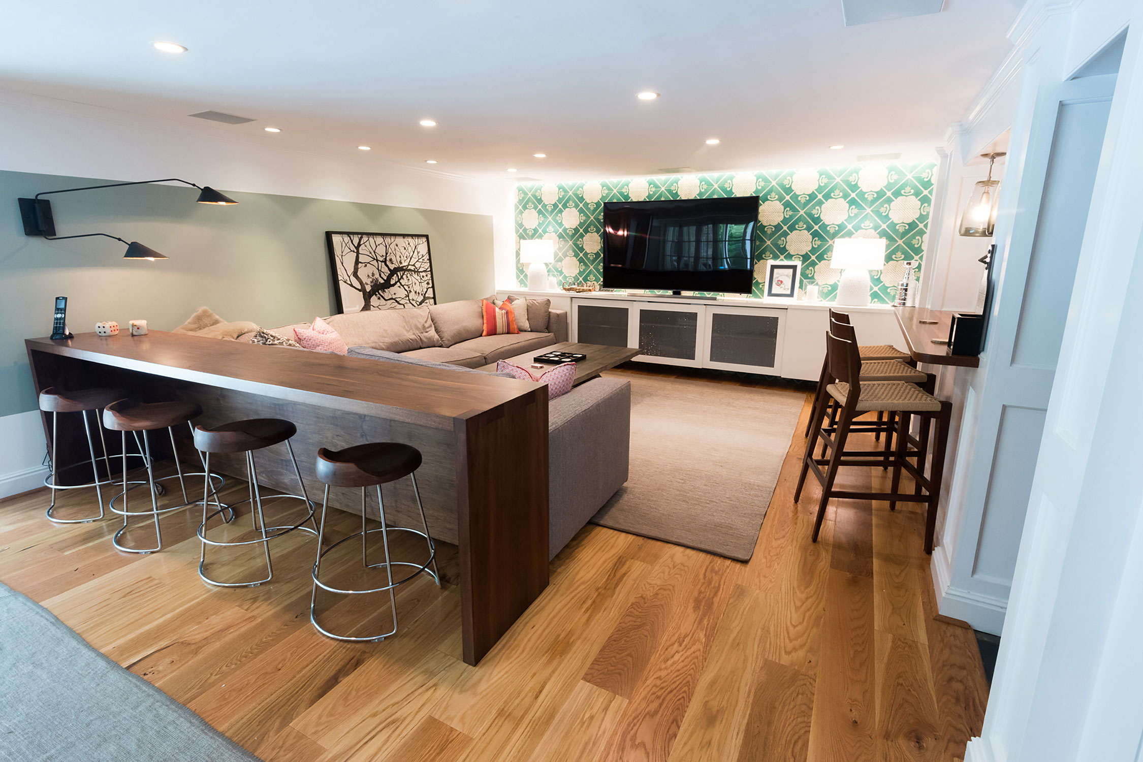 Cozy living area with a sectional sofa, wooden counter with stools, and a large TV. Features a green patterned wall, warm lighting, and stylish decor.