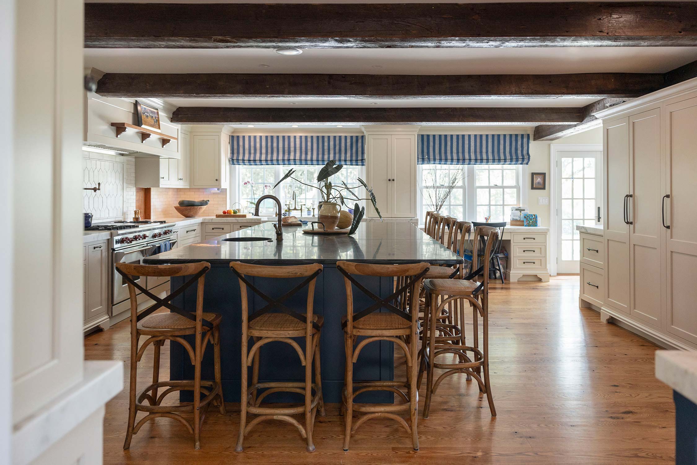 Spacious kitchen with blue island, wooden chairs, and white cabinets. Features striped curtains, wood beams, and large windows over wood floor.