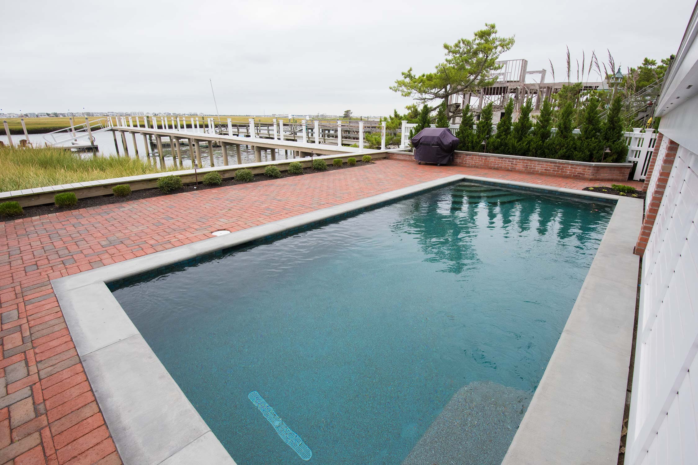 A backyard with a rectangular pool, brick patio, covered grill, and a wooden dock leading to a marshy waterway.