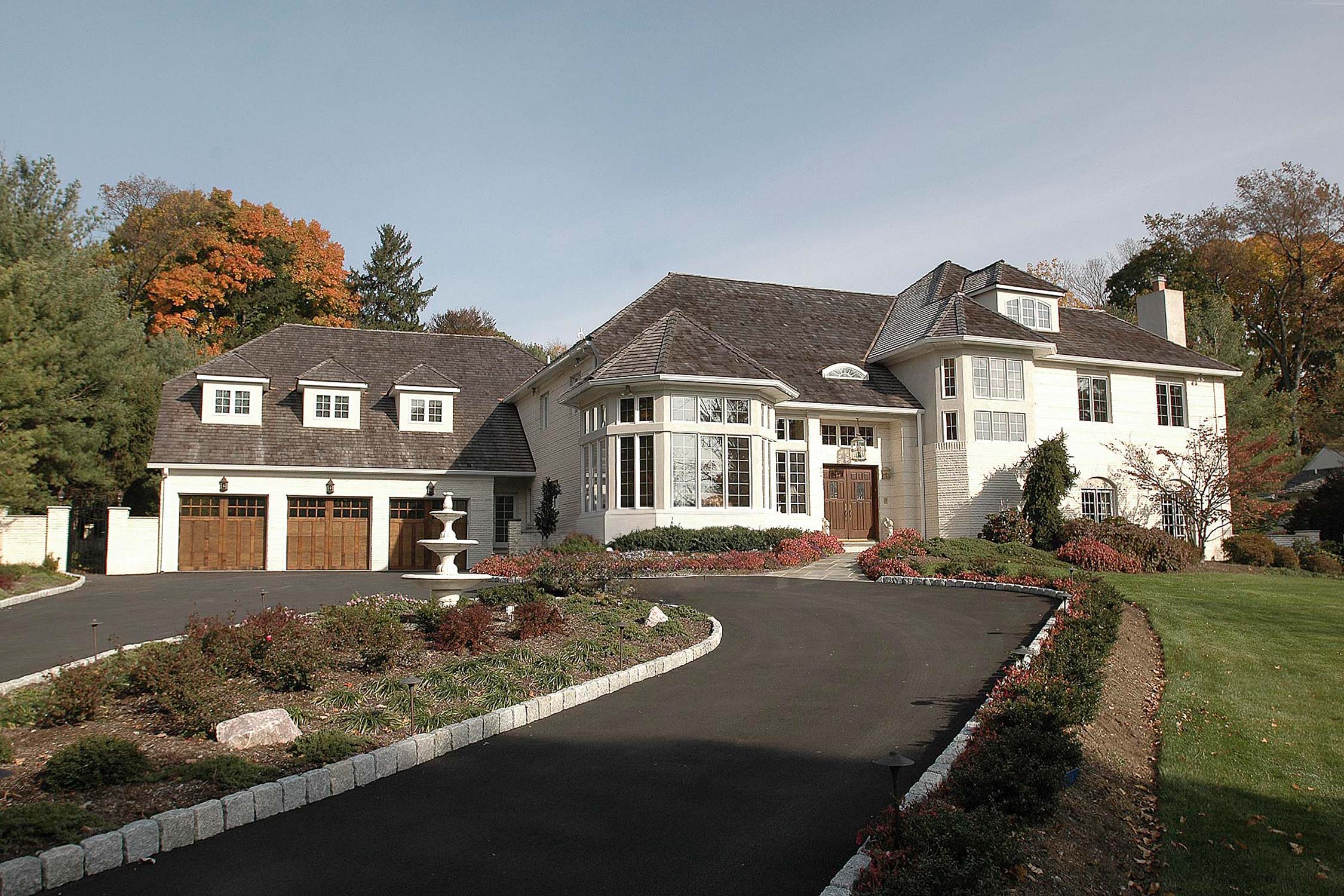 Elegant white home with a curved driveway, double garage, and lush autumn landscaping, featuring a fountain and large bay windows.