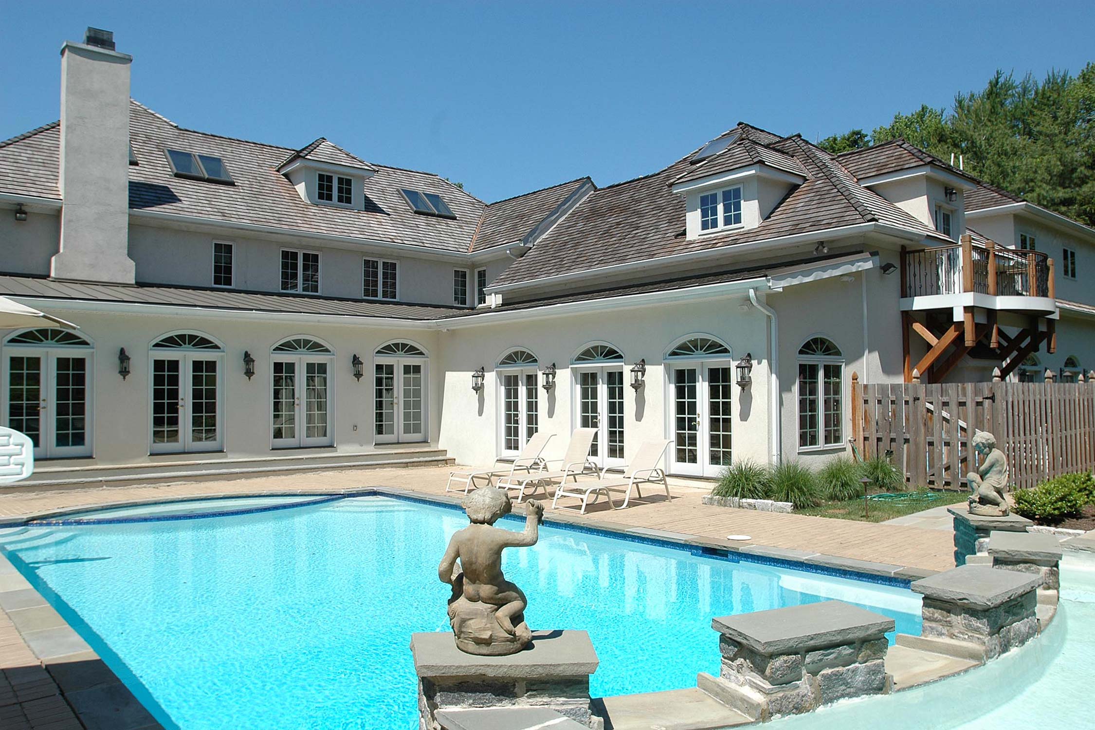 Luxurious home with a sparkling pool, adorned with statues, and a wooden balcony, surrounded by elegant landscaping.