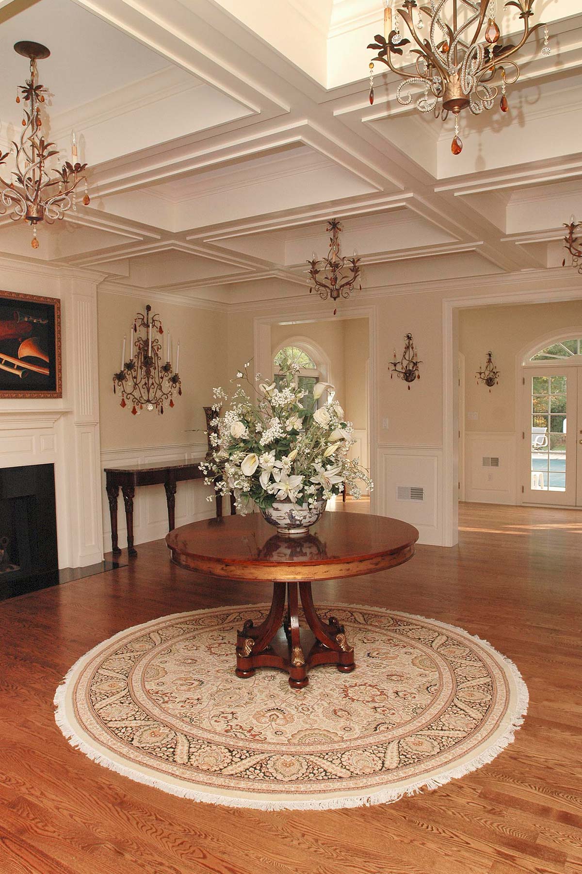 Elegant foyer with a polished wooden table, ornate chandelier, floral centerpiece, and a cozy fireplace.