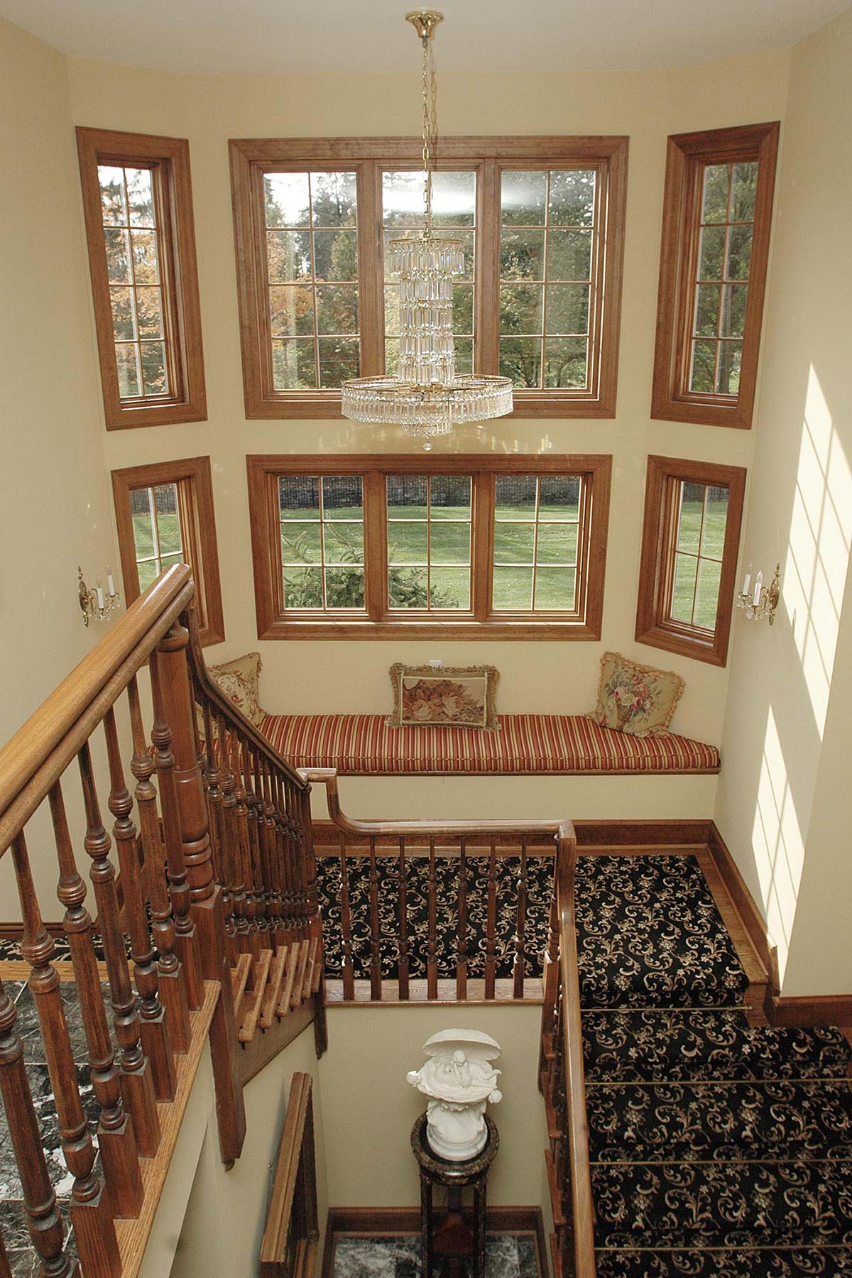 Bright stairwell with large windows, elegant chandelier, cushioned bench, and a marble statue, offering a scenic outdoor view.