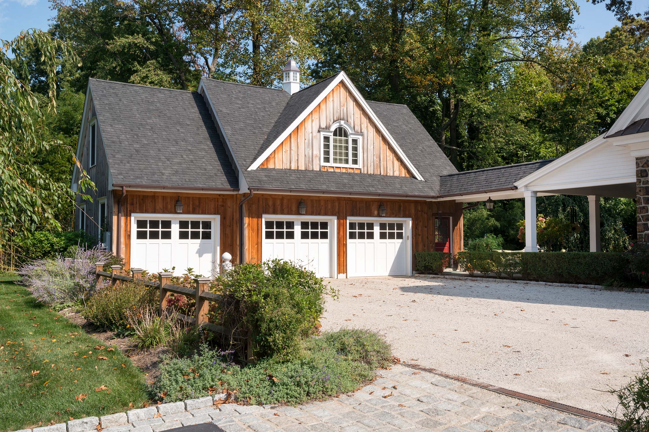 Charming garage with rustic wooden exterior, white doors, and dormer window, nestled among lush greenery, trees, and a paved driveway.