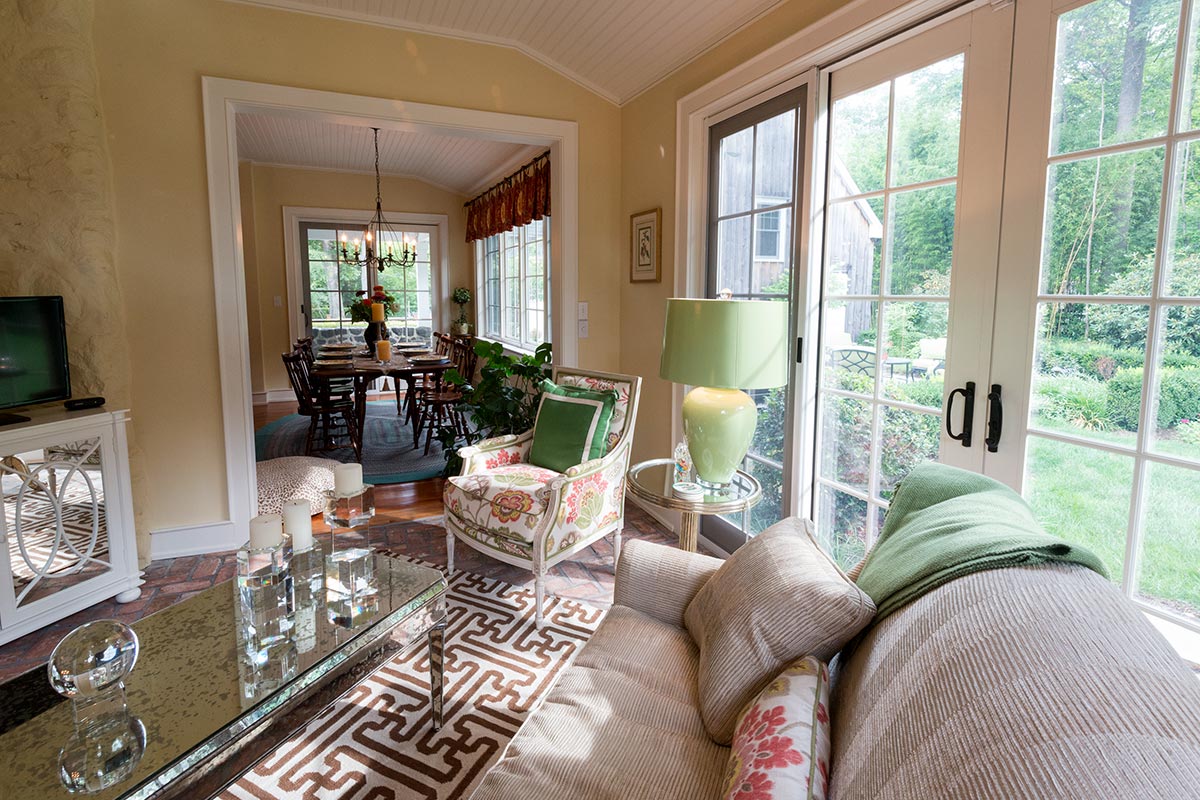 Cozy living room with floral chair, beige sofa, and glass coffee table, featuring large windows with garden views and an adjacent dining area.