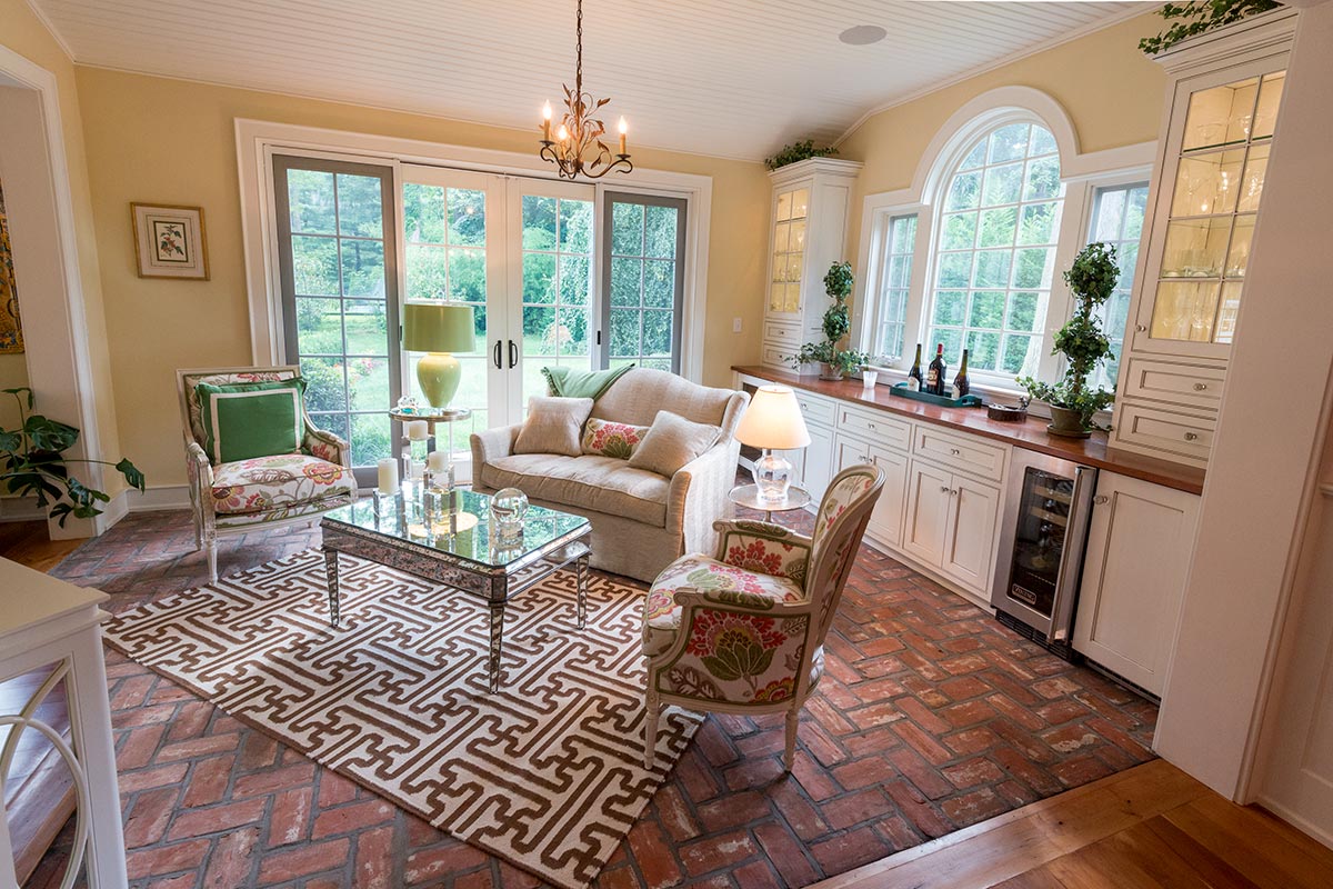 Inviting sunroom with beige sofa, floral armchairs, geometric rug on brick floor, chandelier, and large windows overlooking greenery. Includes built-in cabinets and wine cooler.
