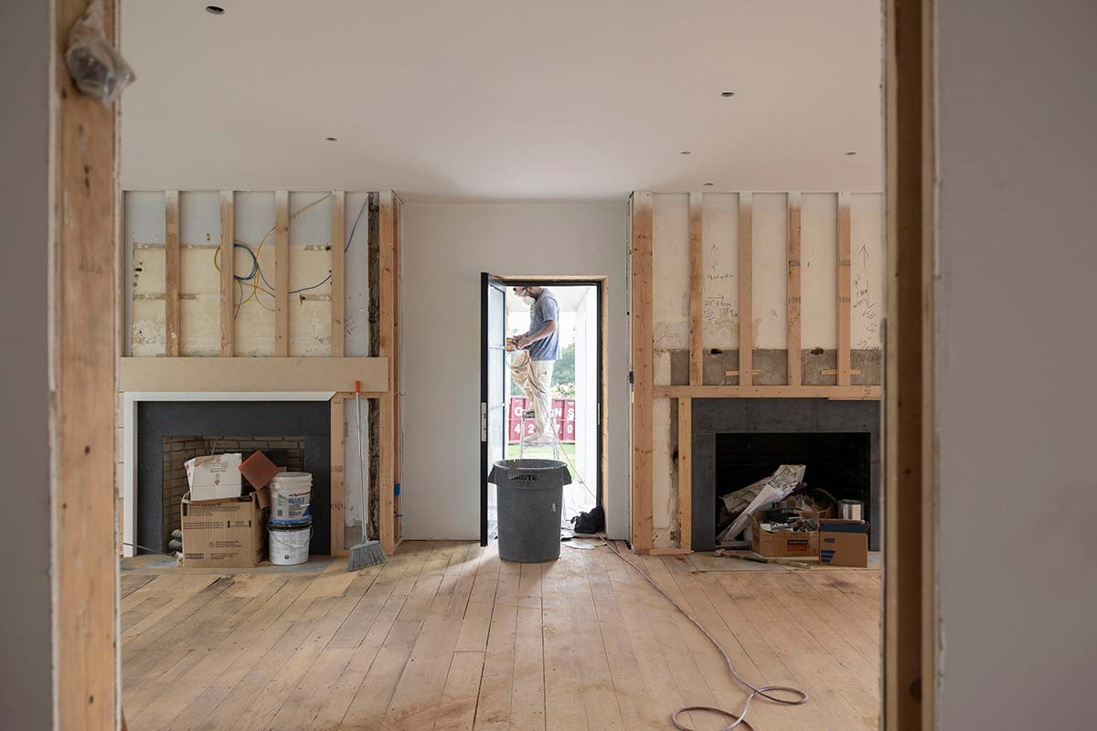 Man on ladder working in an interior room with exposed walls, window, and construction materials, lit by natural light.