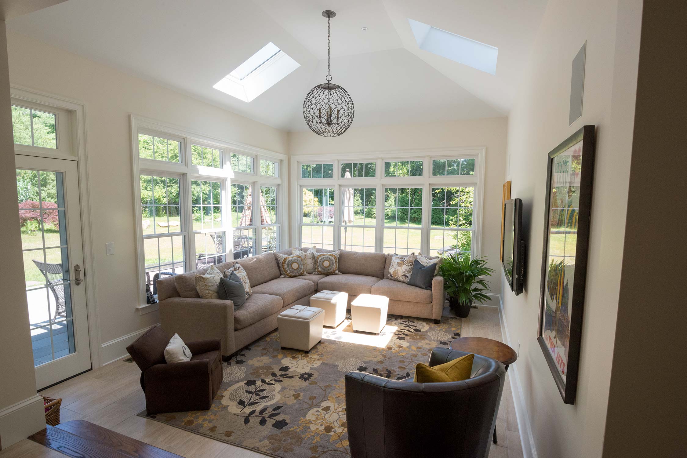 Bright sunroom with large windows, skylights, and a cozy sectional sofa, accented by a floral rug, ottomans, and elegant chandelier.