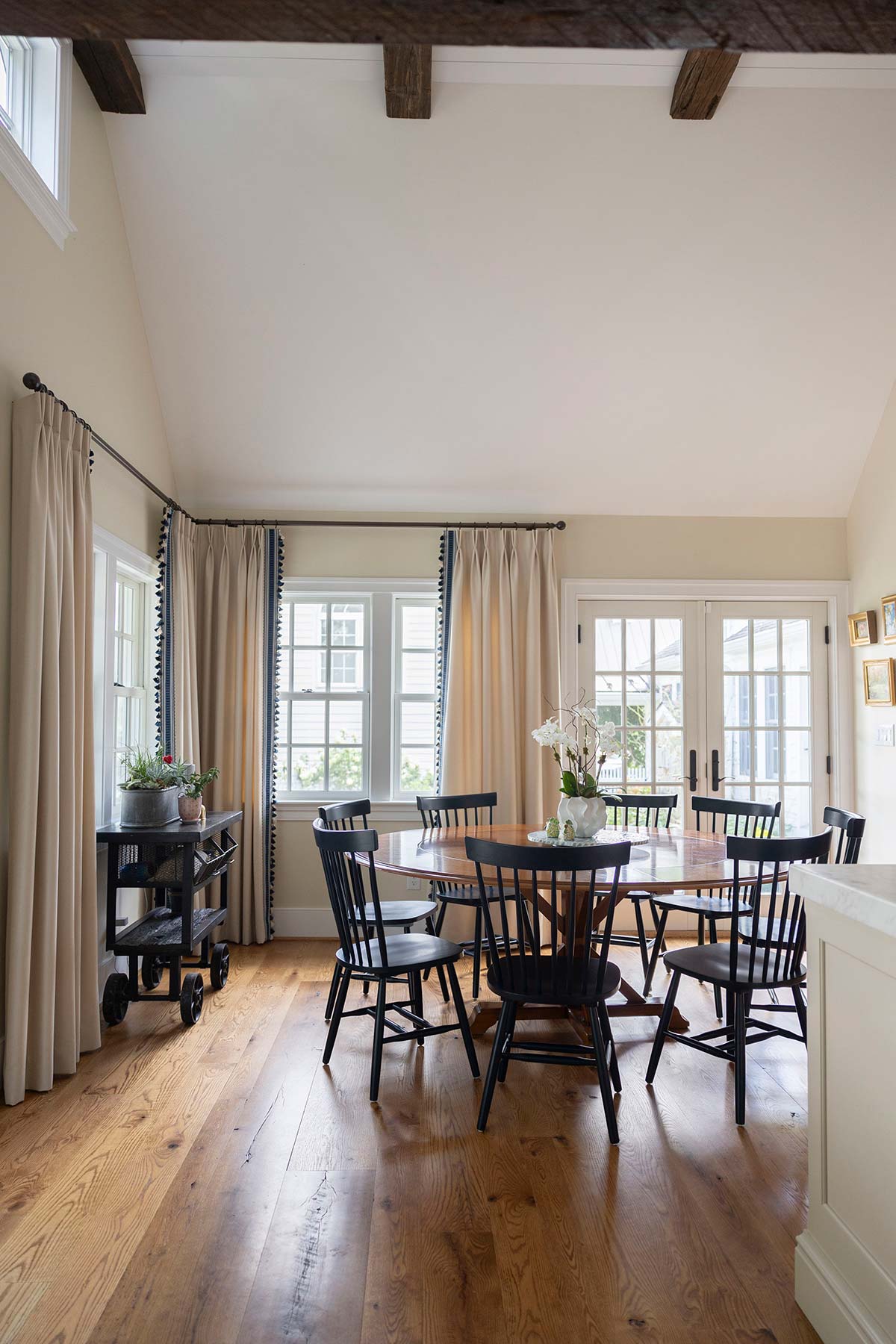 Bright dining area with a round wooden table, black chairs, and large windows with elegant curtains, featuring wooden beams and flooring.