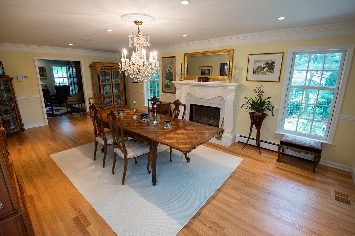 Elegant dining room with crystal chandelier, wooden table, and fireplace. Features hardwood floors, large windows, ornate mirror, and antique furniture.