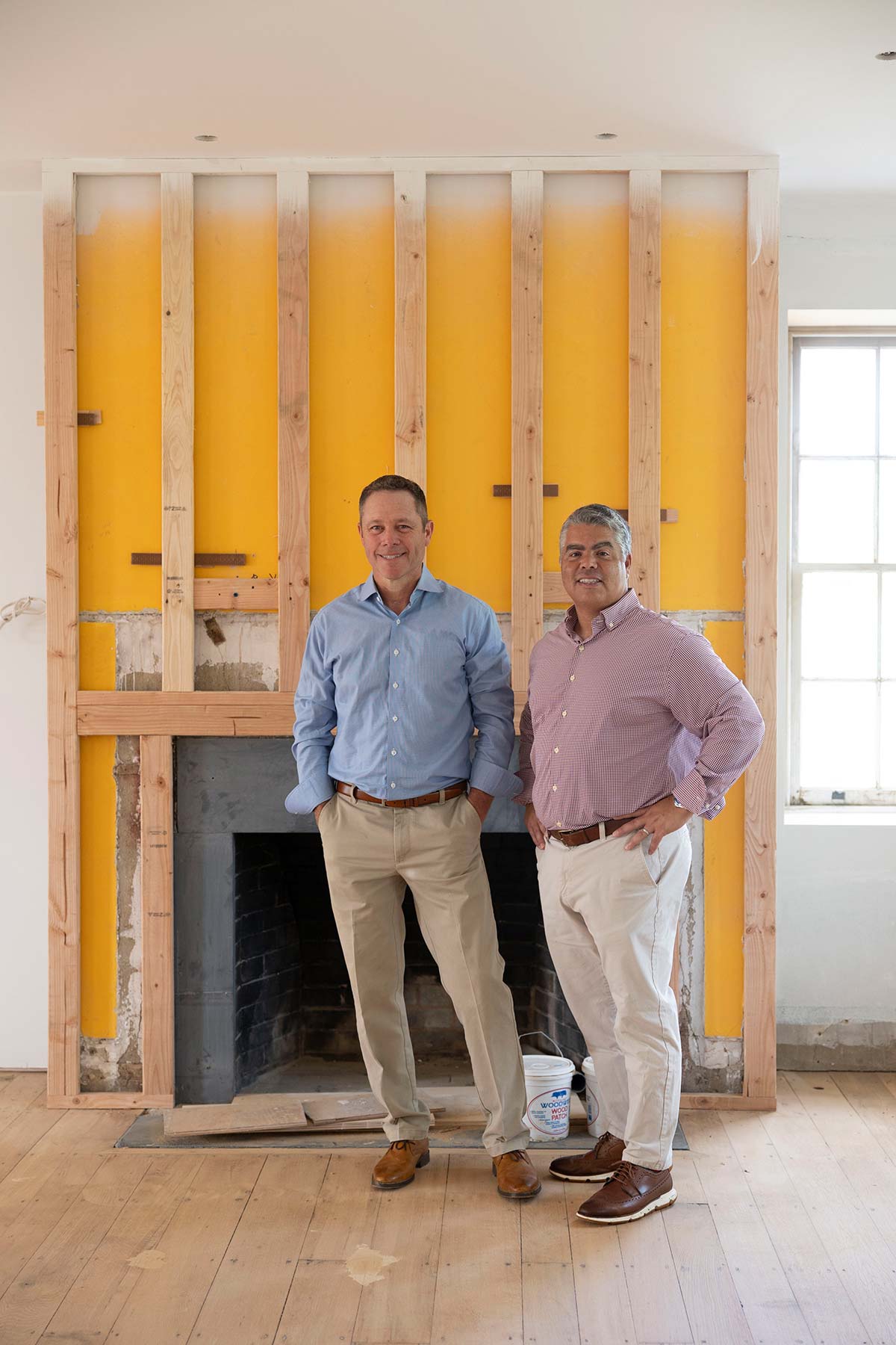 Andy Kerns and Greg Ranieri, Ranieri & Kerns founders, standing in front of a fireplace under construction in a room with wooden floors and white walls.