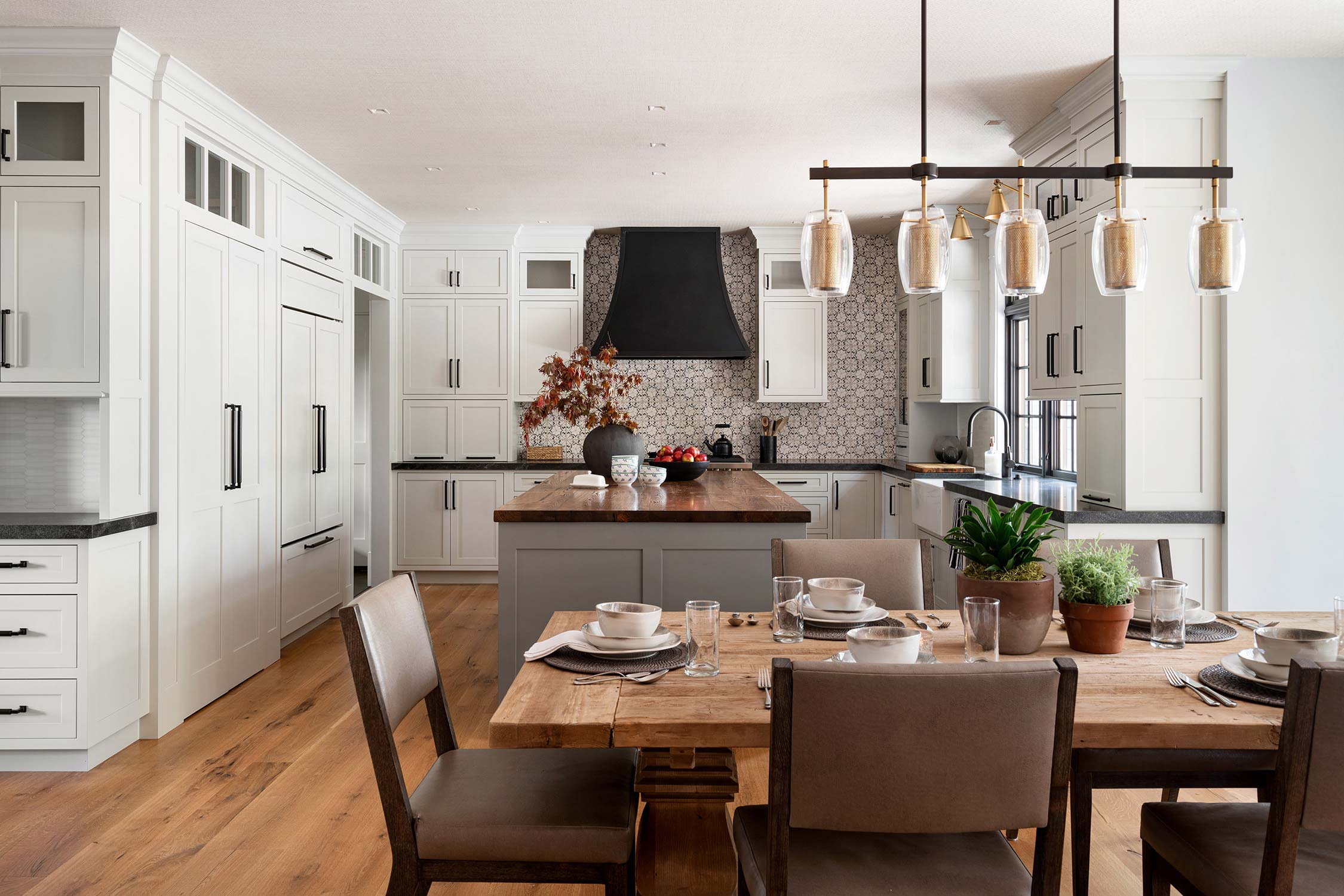 Modern kitchen with white cabinets, gray island, wooden dining table, and pendant lights, featuring plants and set table for a meal.