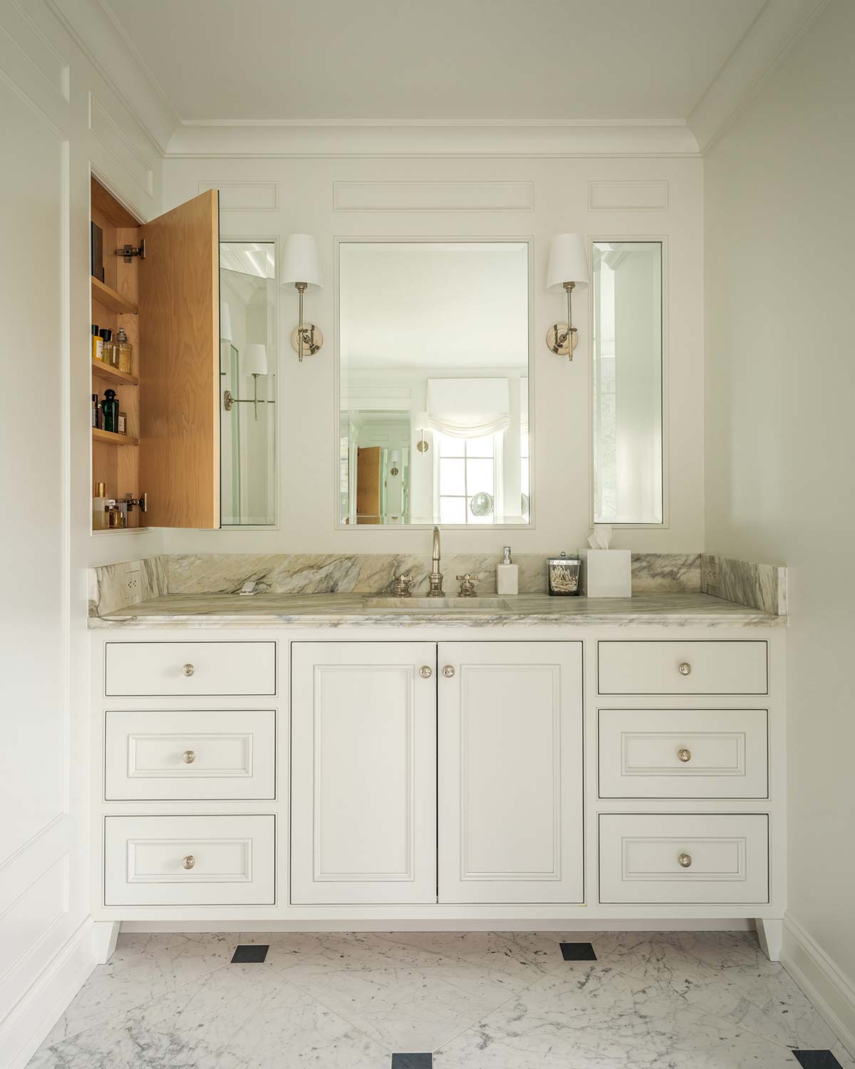 Elegant bathroom vanity with white cabinets, marble countertop, side mirror hiding a medicine cabinet, wall sconces, and patterned marble floor.