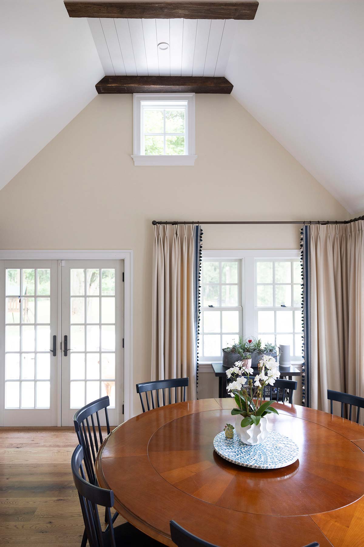 Bright dining room with a round wooden table, black chairs, and floral centerpiece, featuring large windows, curtains, and wooden beams.
