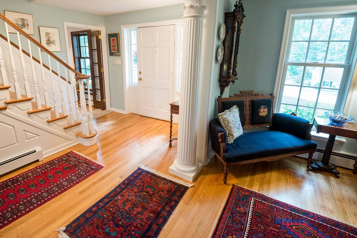Inviting foyer with wooden staircase, ornate column, and blue chaise lounge. Features hardwood floors, large windows, antique clock, and colorful rugs.
