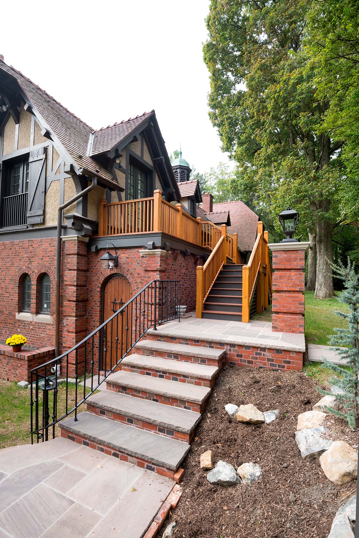 A brick house with a wooden door, balcony, and brick steps leading up, surrounded by trees and landscaping.
